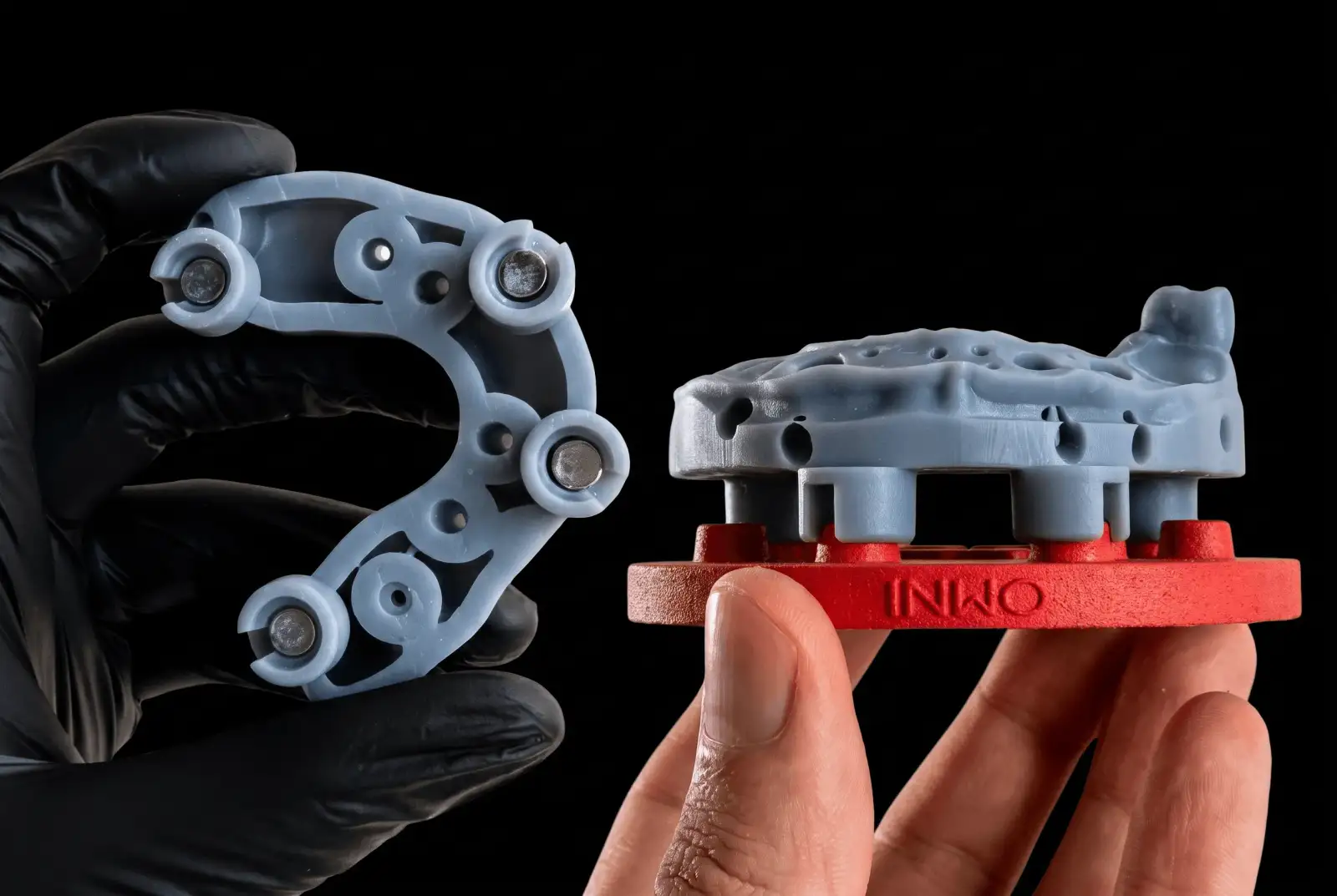 A gloved hand holding the underside of a printed full-arch base with a magnet set in each of its four attachments, beside a printed implant model docked on a red Omni plate